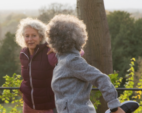 Two women wearing exercise attire, stretching and talking outdoors in a park.
